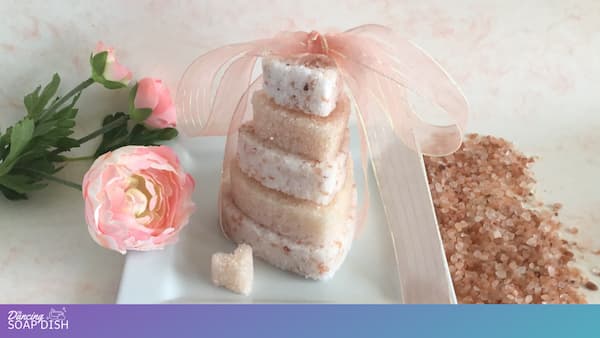 A stack of heart shaped pink Himalayan bath salt cakes tied with a sheer pink ribbon sits on a white square dish. Beside it are a small matching heart-shaped bath salt gem, a pink rose, green leaves, and a pile of pink Himalayan salt crystals.