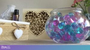 A clear glass bowl holds translucent blue and pink water beads. In the background is a decorative shelf display featuring a wooden box with a white heart ornament tied with twine, a small essential oil bottle, and a textured white vase. In the centre sits a wooden plaque with a large heart cutout filled with smaller heart shapes. Pink flowers in a vase add a soft, cozy touch to the background.