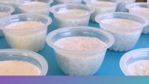 Rows of small clear plastic fruit cups sit on a bright blue surface, each filled with bath bomb mixture speckled with coloured Epsom salt sprinkles. The cups are neatly arranged, creating a uniform display that resembles prepared food samples or a smooth dessert.