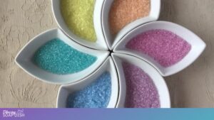 ix white leaf shaped bowls are arranged in a circular flower like pattern, each filled with pastel coloured Epsom salts in yellow, orange, pink, purple, blue, and turquoise. They sit on a textured beige surface, creating a soft, symmetrical, and visually calming display.