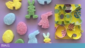 Pastel Easter themed bath salt cakes made from Epsom salts — bunnies, chicks, and eggs in soft yellow, pink, blue, and green — are arranged on the left. On the right, a packaged set labelled “Easter 4pk Cookie Cutters” displays four coloured metal cutters shaped like an egg, a chick, and two bunnies against a yellow decorative background.