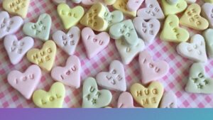 Pastel candy hearts are scattered across a pink‑and‑white checkered surface. Each heart features a short embossed message—such as “LOVE,” “HUG ME,” “LOL,” “I ♥ U,” “UR A ★,” and “B MINE”—in soft shades of yellow, pink, white, and lavender, creating a playful Valentine’s‑themed scene.
