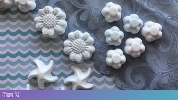 Several pale blue moulded shower melts shaped like flowers and starfish are arranged on a patterned surface. The background includes wavy blue and white designs and blue fabric with swirling motifs.