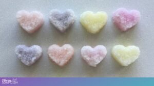 Eight small pastel heart shaped bath salt gems are arranged in two neat rows on a light surface. Each heart has a textured, crystal like finish in shades of pink, purple, yellow, and peach.