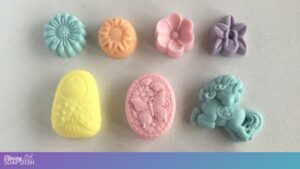 Seven pastel decorative soaps sit on a light surface. The top row features four flower shaped soaps in blue, orange, pink, and purple. Below them are three uniquely shaped soaps: a yellow matryoshka style figure, a pink oval with a butterfly and floral motif, and a blue pony shaped soap.