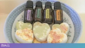A small floral patterned bowl holds four white heart shaped shower melts with soft green and orange patches. Nestled beside them are four essential oil bottles labelled Serenity, Balance, Elevation, and Citrus Bliss. The bowl rests on a plush blue surface, creating a cozy, spa like scene.