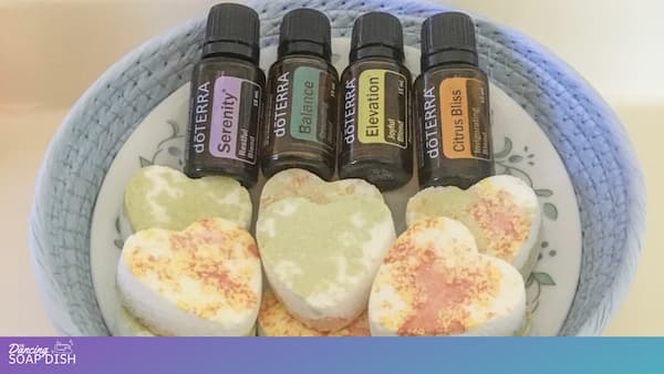 A small floral patterned bowl holds four white heart shaped shower melts with soft green and orange patches. Nestled beside them are four essential oil bottles labelled Serenity, Balance, Elevation, and Citrus Bliss. The bowl rests on a plush blue surface, creating a cozy, spa like scene.