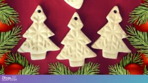 Three white Christmas‑tree‑shaped ornaments with embossed snowflake patterns are arranged on a red background, surrounded by green pine branches and red baubles, creating a festive holiday scene.