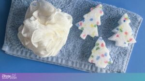 A light blue towel rests on a blue surface, topped with a white bath pouf and three white tree shaped Epsom salt cakes. Each cake contains colourful rock salt pieces in red, green, blue, and yellow, creating a festive, wintery bath display.