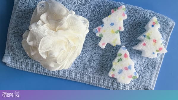 A light blue towel rests on a blue surface, topped with a white bath pouf and three white tree shaped Epsom salt cakes. Each cake contains colourful rock salt pieces in red, green, blue, and yellow, creating a festive, wintery bath display.