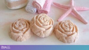Three rose shaped bath salt cakes made from pink Himalayan and Epsom salts sit on a light surface, with rolled white and pink towels behind them. A glass jar filled with layered bath salts and a pink starfish complete the soft, spa themed arrangement.