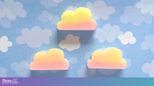 Three pastel cloud shaped soaps with a yellow to pink gradient emulating a sunset rest on a blue background patterned with white illustrated clouds, creating a soft and whimsical sky themed display.