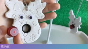 A hand holds a white reindeer shaped ornament with embossed holiday patterns, googly eyes, and a black essential oil vial lid for the nose. The opposite hand is positioning a small red, glass, 1-dram essential oil vial near the nose area, to show that it screws into the ornament to become a red nose. In the background, another similar ornament hangs from a white stand against a green backdrop.