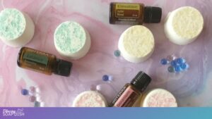 Several round pastel bath bombs with green, yellow, and pink Epsom salt sprinkles are arranged on a marbled pink and white surface. Small blue, purple, and clear water beads are scattered among them, alongside three amber doTERRA essential oil bottles labelled Balance, Geranium, and Elevation.