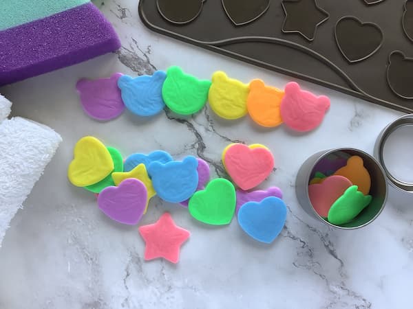 rainbow coloured super thin soap shaped like hearts, starts, and teddies laid out with a silicone macaron mat and a round tin full of single use soap