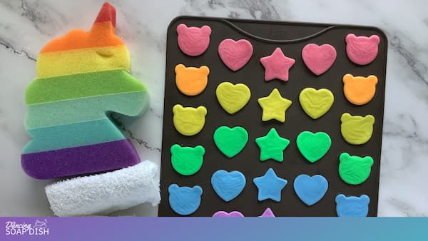 A rainbow striped unicorn shaped sponge sits on the left, featuring horizontal bands of red, orange, yellow, green, blue, and purple. To the right, a silicone macaron mat holds small, flat, brightly coloured soaps in playful bear, heart, and star shapes in shades of pink, yellow, green, blue, and orange, all arranged on a white marble surface.