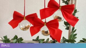 Three clear spherical ornaments hang from red strings, each topped with a large red bow and gold band. Inside the transparent baubles are small pieces of frankincense and myrrh resin, and gold leaf. They’re suspended from a white frame wrapped in green leafy garland, creating a festive holiday display.