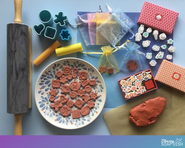 a lump of terracotta clay, mini stamps and cookie cutters surrounding a plate with stamped terracotta charms on it