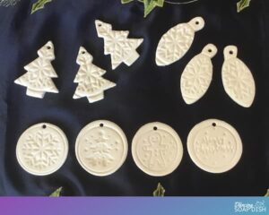 white clay essential oil diffuser ornaments made with christmas cookie cutters and stamps laid out on a navy blue table runner with metallic christmas embroidery