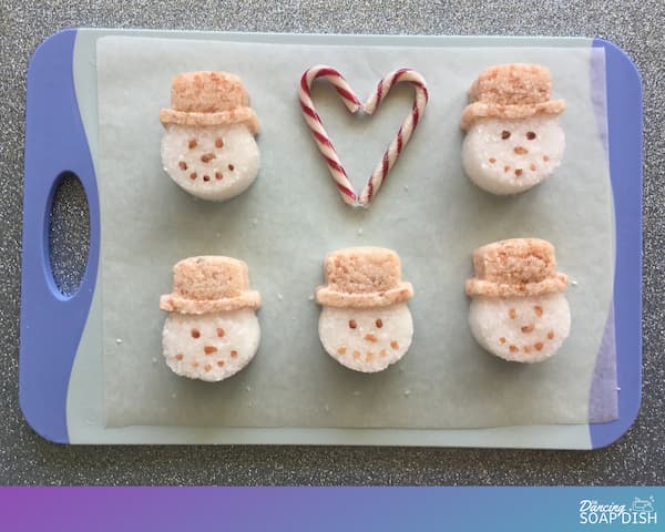 five snowman bath salt cakes laid out on a blue board with two candy canes positioned in the shape of heart