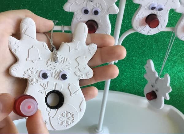 A hand holds a white reindeer shaped ornament with embossed holiday patterns, googly eyes, and a black essential oil vial lid for the nose. The opposite hand is positioning a small red, glass, 1-dram essential oil vial near the nose area, to show that it screws into the ornament to become a red nose. In the background, another similar ornament hangs from a white stand against a green backdrop.
