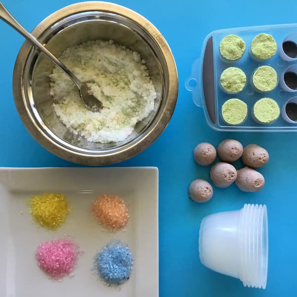 horizontal layout of bath bomb ingredients, colored epsom salt, empty plastic fruit cups and bath bomb inserts