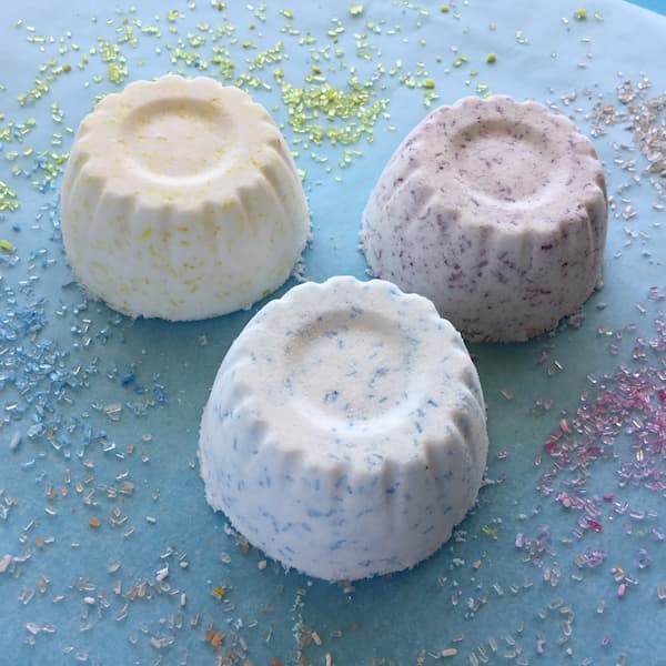 fruit cup bath bombs sitting on a blue background with colored epsom salts scattered around