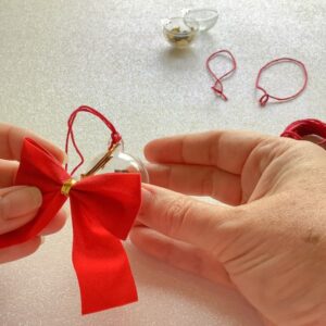 a pair of hands pushing a red velvet bow through the hole of a plastic bauble
