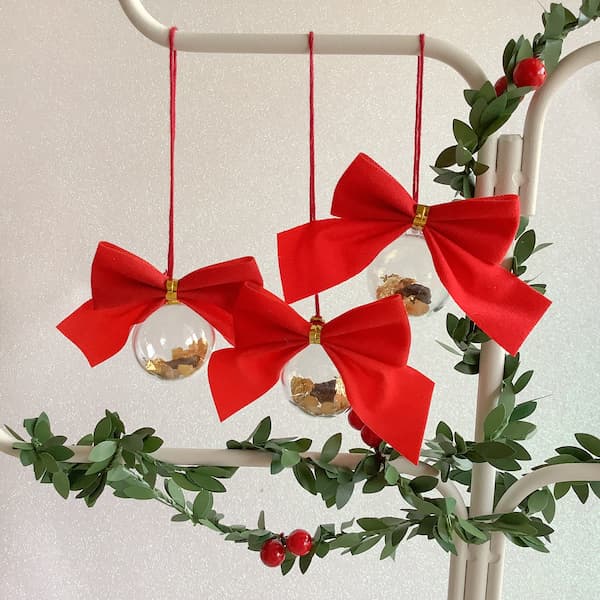 three clear baubles with red bows and gold, frankincense and myrrh pieces inside hanging from an ornament stand covered with fake holly