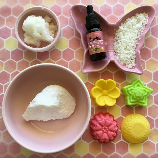 an overhead view of shea butter, coconut oil, beeswax and rosehip oil laid out in assorted pink bowls with four floral silicone molds