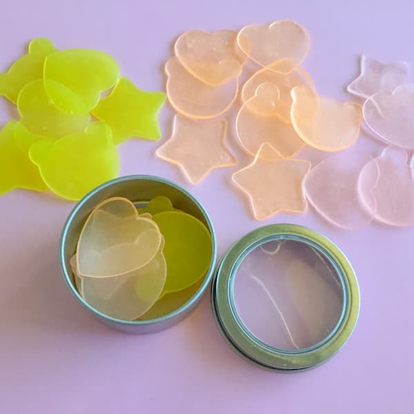 yellow, orange and pink super thin soap slivers in the shape of stars, hearts and teddy bear heads sitting in, and scattered around, a round metal craft tin