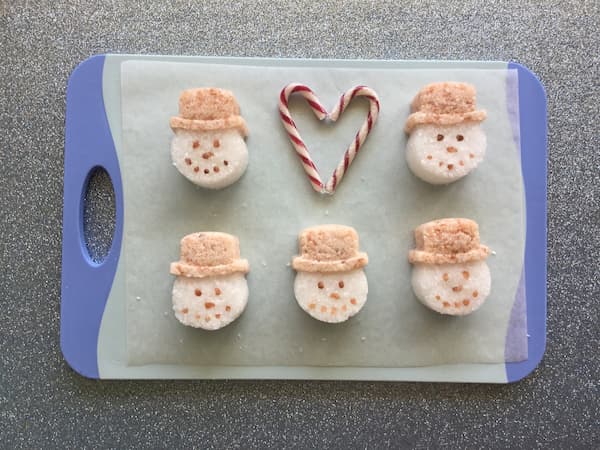 five snowman bath salt cakes laid out on a blue board with two candy canes positioned in the shape of heart