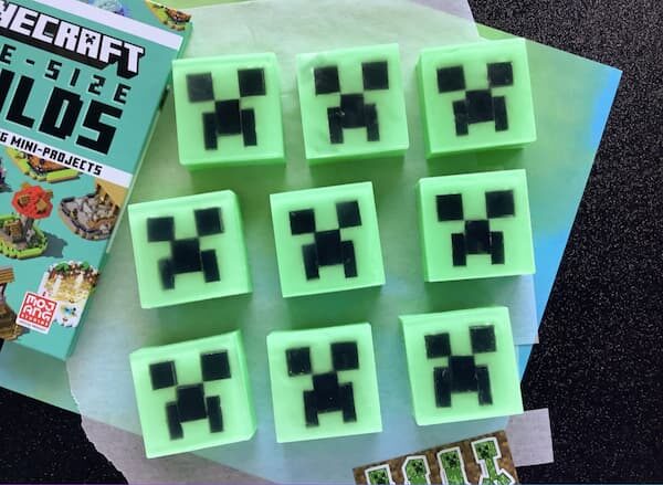Nine square green soaps are arranged in three rows, each featuring a black pixelated Creeper face inspired by Minecraft. A partially visible Minecraft book sits to the left, and the soaps rest on a light green surface.