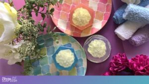TThree hexagonal soaps with white carnation soap embeds sit on colourful patterned plates. Each soap has a different coloured background of orange, purple or blue. Rolled towels in blue, white, and lavender surround the setup, along with scattered flowers and greenery, all arranged on a pink surface for a coordinated, spa style display.