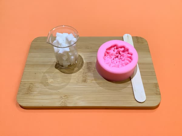 a 50ml glass beaker filled with small cubes of white melt and pour soap base sitting on a bamboo board next to a carnation soap mould and a wooden popsicle stick