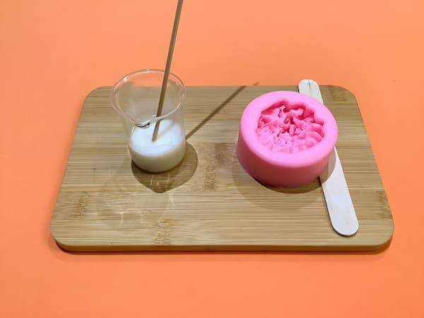 a 50ml glass beaker filled with melted white soap base and a bamboo skewer sitting on a bamboo board next to a carnation soap mould and a wooden popsicle stick