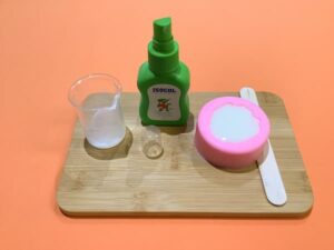 an empty glass beaker sitting on a bamboo board next to a bottle of isopropyl alcohol and a carnation silicone mould full of white soap
