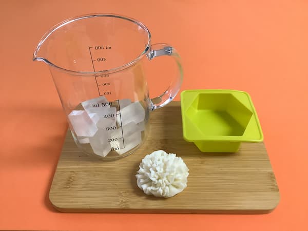 a glass measuring jug filled with cubes of clear melt and pour soap base sitting on a bamboo board next to a hexagonal silicone mould and a white soap carnation