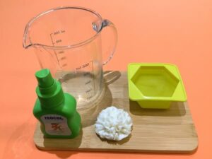 a glass measuring jug filled with melted clear melt and pour soap base sitting on a bamboo board next to a hexagonal silicone mould, a bottle of isopropyl alcohol, and a white soap carnation