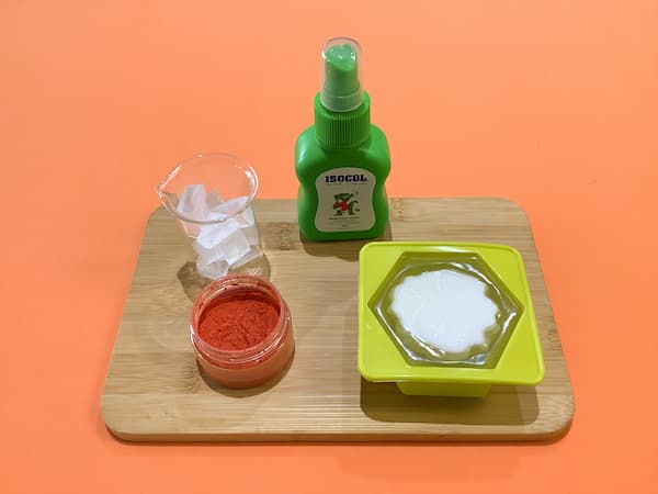 a small glass beaker of clear melt and pour soap base sitting on a bamboo board with a container of orange mica, a bottle of isopropyl alcohol, and a hexagonal soap mould filled with clear melt and pour soap base with an upside down white carnation soap positioned in the middle