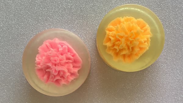 two round soap bars with a carnation embedded in the centre, one pink, one orange.