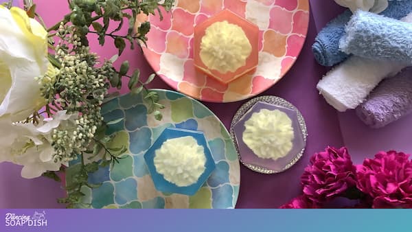 TThree hexagonal soaps with white carnation soap embeds sit on colourful patterned plates. Each soap has a different coloured background of orange, purple or blue. Rolled towels in blue, white, and lavender surround the setup, along with scattered flowers and greenery, all arranged on a pink surface for a coordinated, spa style display.