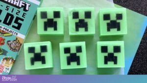 Six square green soaps are arranged in two rows, each featuring a black pixelated Creeper face inspired by Minecraft. A partially visible Minecraft book sits to the left, and the soaps rest on a light green surface.