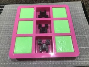 a nine-cavity square soap mould, six cavities of which are filled with green soap, three of which are filled with black minecraft creeper faces
