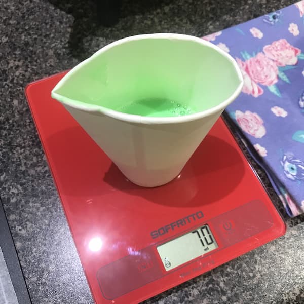 melted green soap base in a paper cup sitting on a set of kitchen scales displaying '70g'
