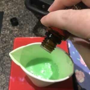 a hand pouring essential oils from a bottle into a paper cup filled with green melt and pour soap