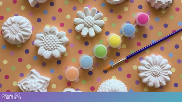 White plaster fridge magnets shaped like flowers and decorative motifs are arranged on an orange polka dot background. Small pastel paint pots—pink, blue, yellow, green, and peach—sit among them, along with a paintbrush, creating a colourful art and craft setup.