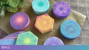 Seven handmade soaps with vibrant pastel tie dye patterns are arranged on a brown cloth. Most are round, with one hexagonal bar, all featuring swirls of blue, purple, pink, yellow, and green. A striped towel sits in the lower left and green plants appear in the upper left.
