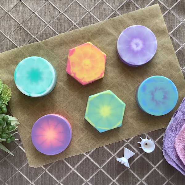 six tie dye melt and pour soap bars laid out on a piece of brown paper with two paint pouring tools