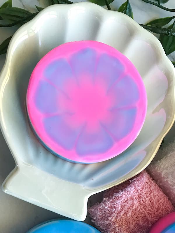 a round soap bar sitting on a clam shell plate. the soap bar has a beautiful tie dye effect coloured pink, blue and a hint of white